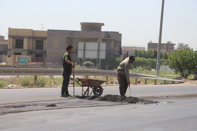 عاملان ينفذان أعمال ترقيع وصيانة الإسفلت يدوياً باستخدام عربة وأدوات تسوية.