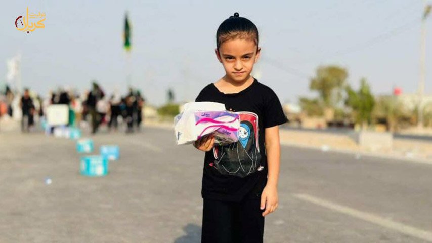 طفلة تقف وسط طريق زوار الأربعينية حاملة علبة مناديل ورقية لتقديم الخدمة.