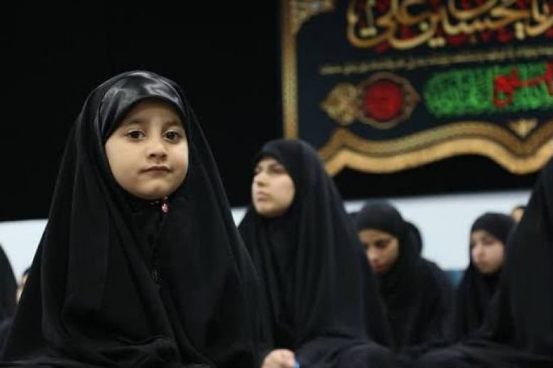 طفلة ترتدي العباءة السوداء تجلس بين جمع من النساء في مجلس حسيني.