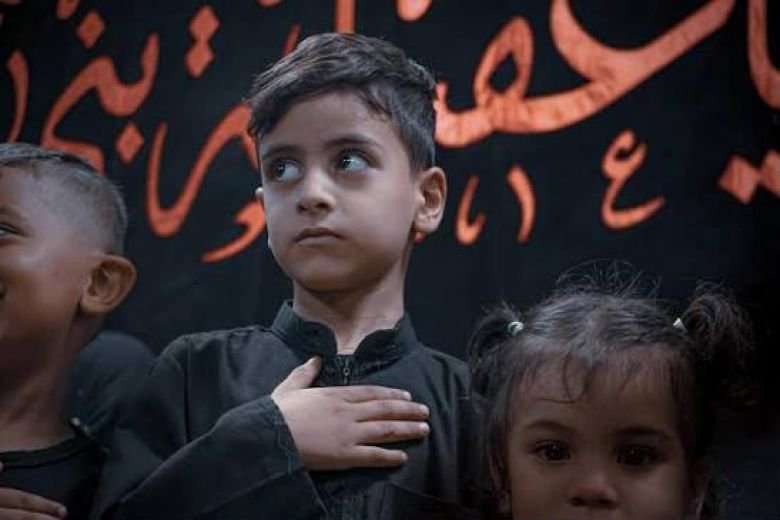 طفل يرتدي قميصاً أسود يضع يده على صدره في مجلس حسيني.