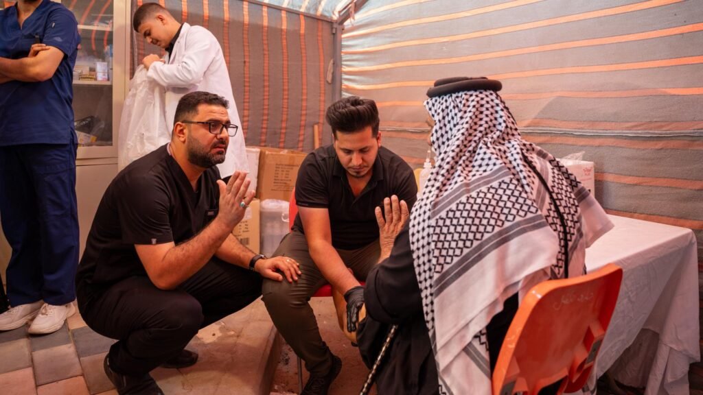 مسعفان يتحدثان مع رجل مسن يرتدي الكوفية ويفحصان قدمه داخل خيمة مفرزة طبية.
