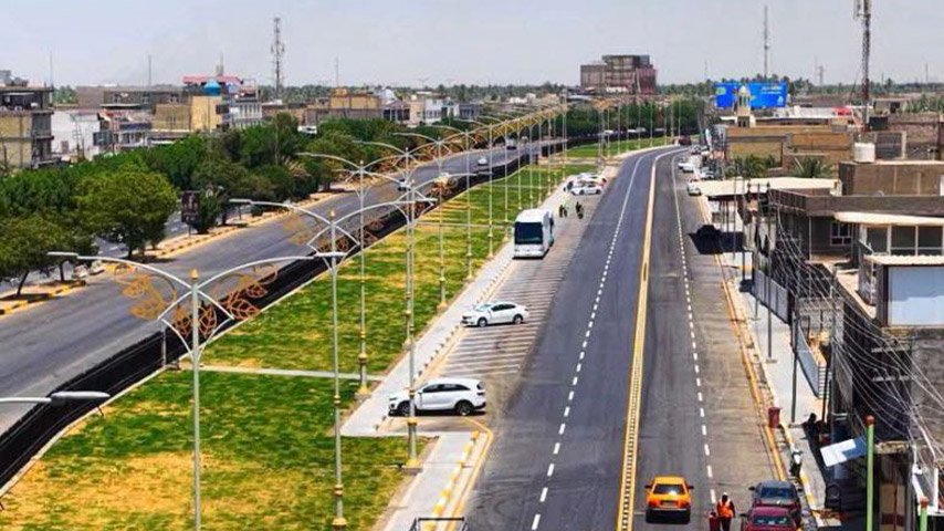 طريق حديث متعدد المسارات مزود بجزيرة وسطية خضراء وأعمدة إنارة ومواقف جانبية للمركبات.