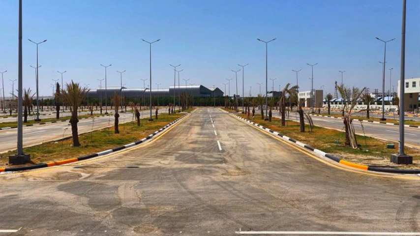 طريق داخلي واسع مزود بأعمدة إنارة ومساحات خضراء وأشجار نخيل ضمن مشروع خدمي حديث.