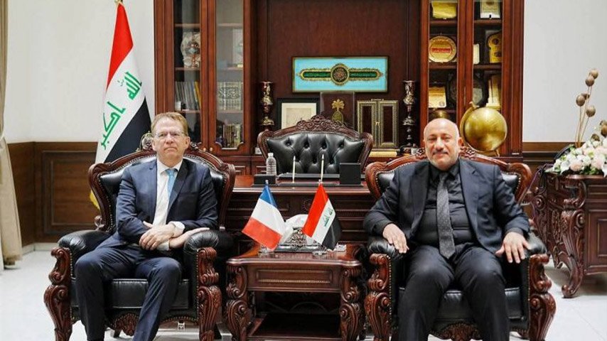 مسؤول عراقي ودبلوماسي أجنبي يجلسان في لقاء رسمي وبجوارهما العلمان العراقي والفرنسي.