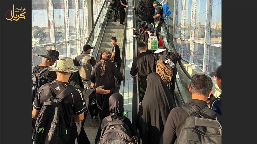زوار يرتدون السواد ويحملون حقائب ظهر أثناء صعودهم جسر مشاة كهربائي مغلق بالزجاج.