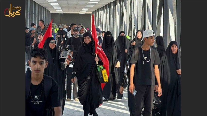 حشد من الزوار يرتدون الملابس السوداء ويحملون رايات حمراء يسيرون داخل جسر مشاة مغلق.