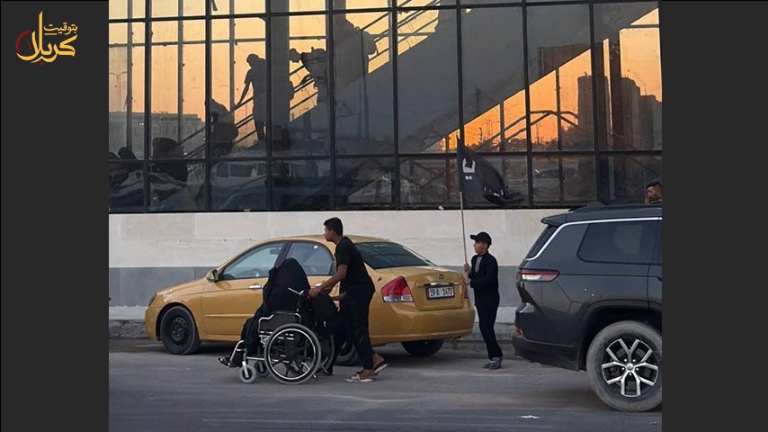 شاب يدفع كرسيًا متحركًا وصبي يحمل راية سوداء قرب سيارة أجرة ومبنى زجاجي.