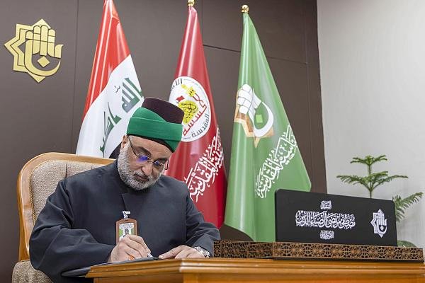 شخصية دينية تكتب في مكتب أمام رايات عراقية ورايات العتبات.