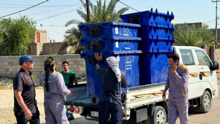 شاحنة محمّلة بحاويات نفايات زرقاء أثناء تفريغها من الكوادر الخدمية.