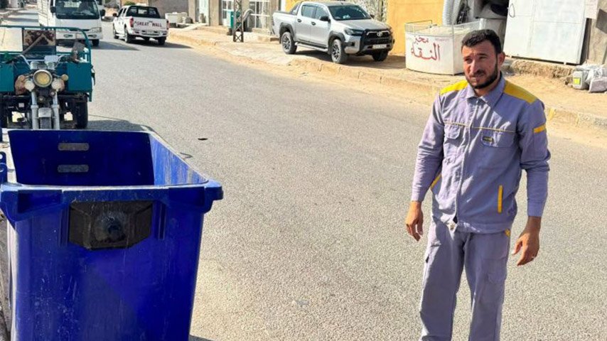 عامل بلدية يقف بجانب حاوية نفايات زرقاء في الشارع.