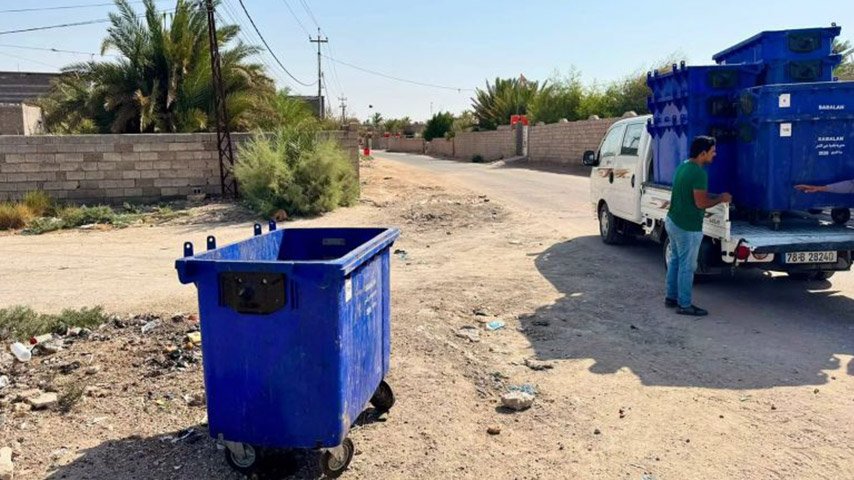 حاوية نفايات زرقاء موضوعة في شارع ترابي بالقرب من شاحنة نقل.