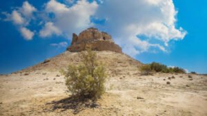 تلة صحراوية تعلوها بقايا حصن أو أطلال أثرية تحت سماء صافية وغيوم بيضاء.