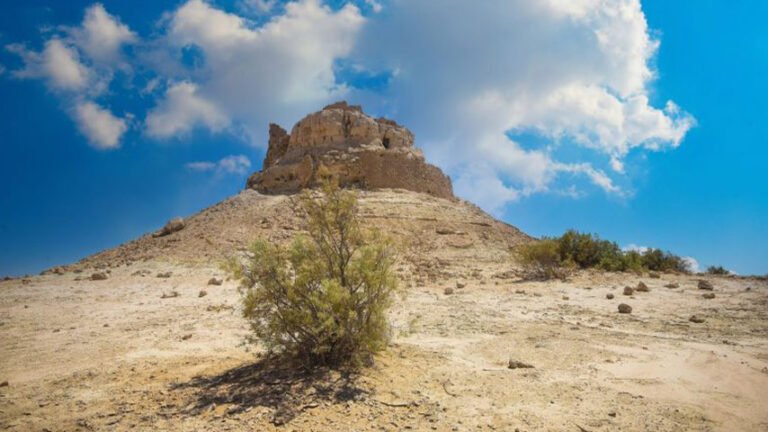 تلة صحراوية تعلوها بقايا حصن أو أطلال أثرية تحت سماء صافية وغيوم بيضاء.