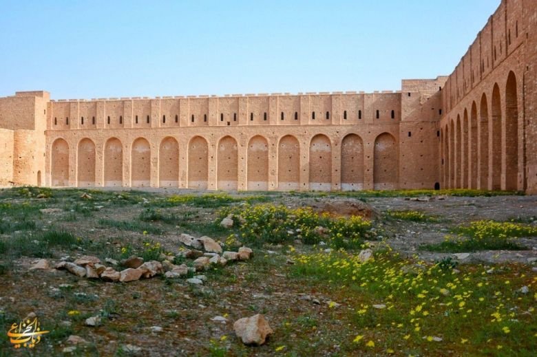 جدار عريض لحصن الأخيضر يحتوي على صف من الأقواس المتتالية أمام ساحة مفتوحة تتناثر فيها الأعشاب والزهور الصفراء.