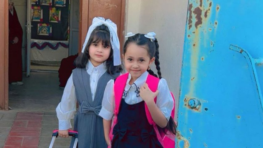 طفلتان ترتديان الزي المدرسي وتملكان حقائب مدرسية تبتسمان أمام مدخل المدرسة.