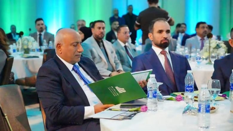 محافظ كربلاء يقرأ مستندات داخل كتيب أخضر أثناء جلوسه في مؤتمر رسمى بجانب شخصيات رسمية.